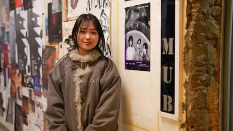 BBC/ Hosu Lee 29-year-old Lee Hae stands in front of a wall covered in posters. She wears a heavy beige coat with fur trim.