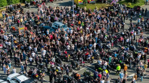 Ross Bisley An aerial image showing hundreds of people gathered around Josh in a car park. The ground is barely visible as the crowd is densely packed together, and there are lots of motorbikes parked in spaces.