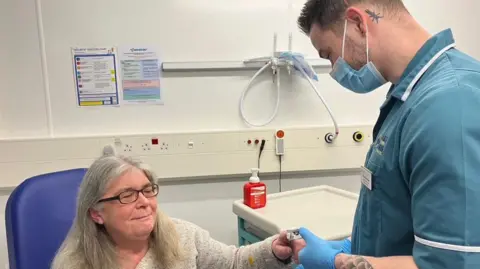 Lesley Jenkins who has long grey hair and glasses, sat on a blue medical chair, wearing a grey jumper. She has her arm outstretched as a medic wearing blue scrubs and a face mask pins a blood pressure monitor to her finger. Medical equipment is visible in the background.