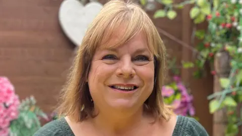 Jayne Biggs A woman who is standing outside in front of a tree and flowers. She is wearing a green top and is looking directly at the camera and smiling.