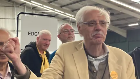 A man with white hair, rimless glasses and a moustache celebrates an election win. He's punching the air with glee, wearing a cream jacket, and a Liberal Democrat orange badge pinned to his left lapel. 