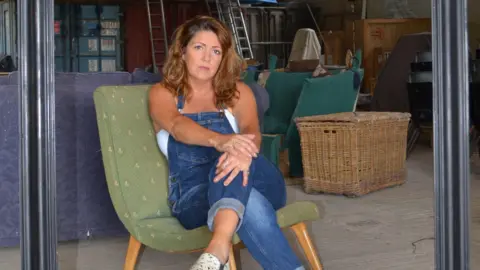 Lianne Morgan A woman sat on a big green chair in blue denim dungarees in a studio. 