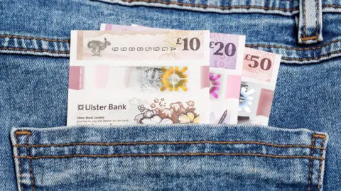 A close-up of three notes in the back pocket of a pair of jeans. The notes are a £50, £20 and £10 from the Ulster Bank.