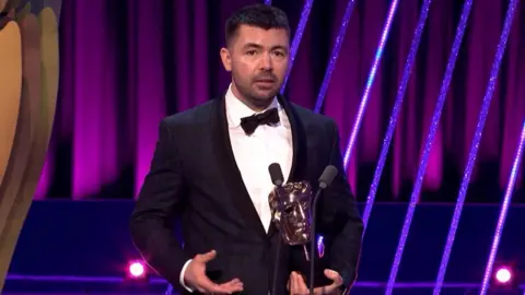 Rich Peppiatt standing on stage in a dark suit and bow tie holding the Bafta award and speaking into microphones. 