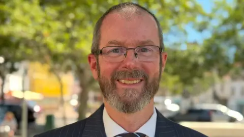 A man wearing glasses, a dark pinstripe suit and a striped tie stands outdoors in a tree-lined area. The image is a head-and-shoulders portrait with a blurred background showing trees, parked vehicles and buildings in daylight.