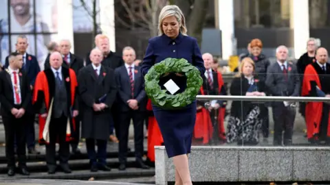 Getty Images Michelle O'Neill in navy skirt suit and black heels, laying a green laurel wreath at remembrance event in Belfast City Hall, out of focus crowd in background watch on in dark clothes