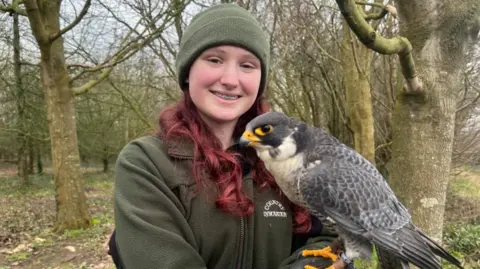 A girl with red hair and braces wearing a green fleece and green hat stands in winter woodland holding a mottled grey and white Peregrine Falcon on her left arm. The bird has orange nostrils and feet and black eyes. It's looking beyond the camera to the left. 