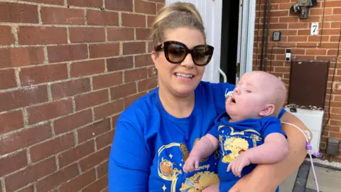 A woman with tied back dark blonde hair and big black sunglasses smiles as she holds her baby boy, who has a feeding tube in his nose. She is standing on her garden patio. Both she and her baby are wearing blue The Lion King T-shirts.