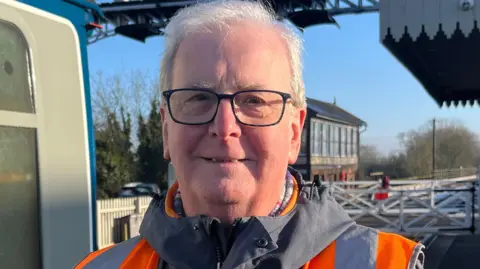 Tom Jackson/BBC Michael Purcell is standing to the side of a train and smiling at the camera. He has grey hair, square framed glasses and is wearing an orange high vis vest. It is blue sky behind him and the light from the sunrise is shining on his face. 