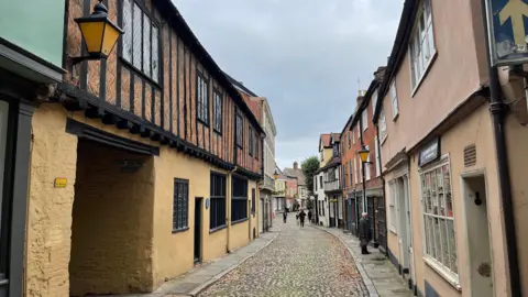 A view down historic Elm Hill in Norwich, a narrow cobbled street. A 16th Century oak-beamed building stands to the left, with black framed latticed windows. Its ground floor plasterwork is painted a sandy colour. The street has a thin path either side of the cobbles, with the old buildings either side creating a continuous terrace which curves to the right in the distance. A man looks in the window of a shop further along the street. 