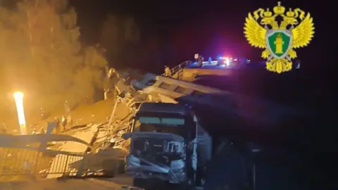 A vehicle surrounded by debris at night. People are standing a short way away watching.
