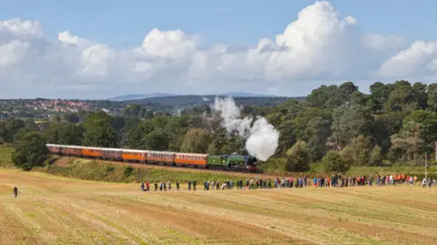 A line of people stand in a field and watch as the iconic Flying Scotsman locomotive travels along a railway in the middle of the countryside. Steam can be seen coming out the front of the train. There are trees on the other side of the track and a settlement is visible on the horizon.  