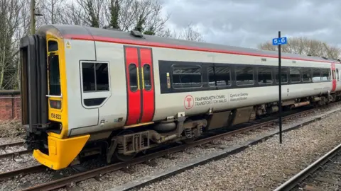 A red, grey and yellow Transport for Wales train on a track.