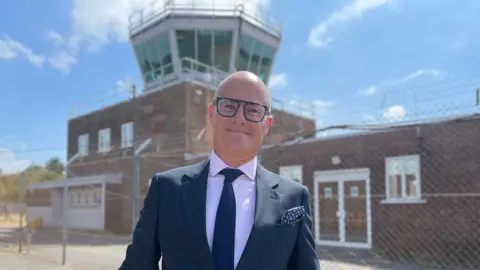 Mark Ansell/BBC A bald man with glasses in a smart black suit and light pink t-shirt smiles in front of the air traffic control tower. 