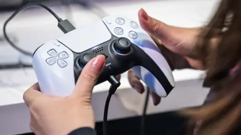 A close up of the Playstation 5 game controller in the hands of a women with pale pink nail varnish. She appeasrs to be at a promotional event for Playstation