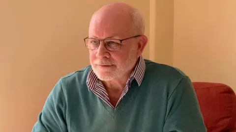 A white man with a white beard and glasses, wearing a green jumper and a blue stripey shirt sits in a red armchair in front of a creme wall. 