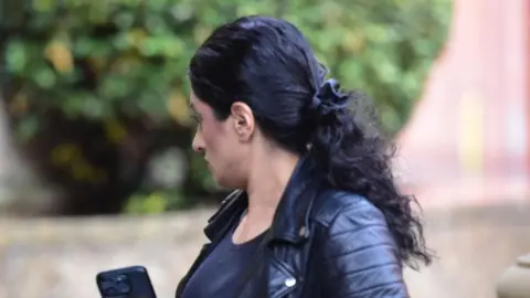 Spindrift A woman with long dark hair and wearing a dark leather jacket walking outside court - she has her head turned away from the camera and a phone in her hands