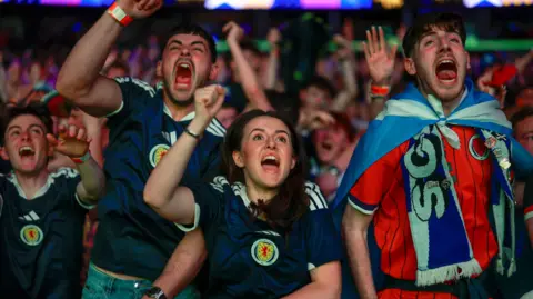 Fans celebrate in the OVO Hydro as Scotland score against Haiti. In the foreground are three men and one woman, all cheering and wearing Scotland strips. Rows of supporters can be seen behind them.