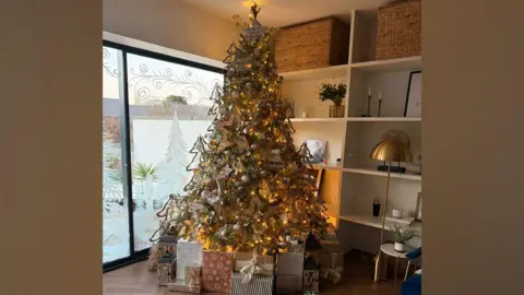 Claire Powell A Christmas tree lit up with large stars and lights. There are lots of beautifully wrapped presents under the tree. There is festive stencilling on some glass doors which lead to the garden behind the tree.