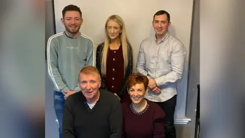 O'Hagan family Family photo. Cahir is wearing jeans and a green jumper, Briege, with long blonde hair is wearing a burgundy dress, beside Paul in a grey shirt. In the foreground, sitting, is Kevin, with ginger hair wearing a dark top and mother Briege in burgundy top.