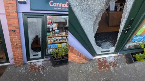 Tom Richards Two pictures in a collage side by side of a smashed window of the Canna Deli store in Pontcanna Mews. The left image shows a large hole in a glass window pane inside a door. Plants can be seen outside the front of the store, and shattered glass covers a doormat and the ground outside of the door. 