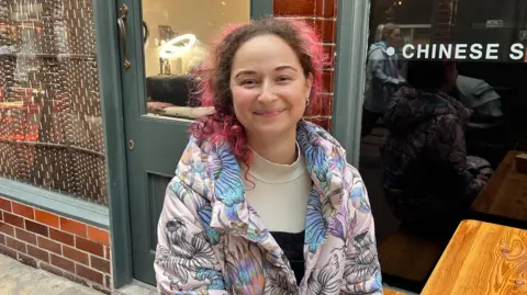 Danielle Harris seated outside one of the food outlets at the arcade. She has red and brown hair and is wearing a multi-coloured jacket. She is smiling at the camera.