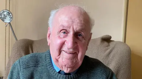 Douglas Cracknell smiles at the camera while he sits in a chair. He wears a green knitted round-necked jumper with a blue shirt underneath, and tie.