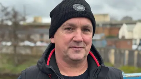 A man wearing a black hat, coat and top. He has stubble and has blue eyes.