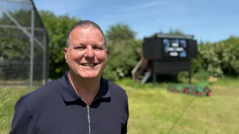 Harvey Burgess stands in front of a cricket net and a black scoreboard, smiling at the camera, dressed in a navy blue shirt. 
