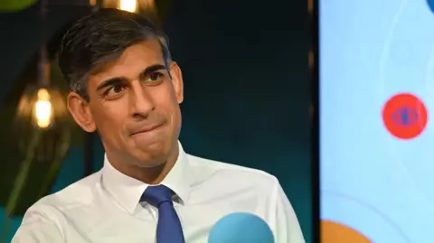 A medium close up shot of Rishi Sunak. He wears a white shirt and a blue tie.
