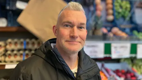 A man smiles into the camera. He has short blonde hair and stubble and wears a black coat. Fruit and vegetables are pictured behind him.