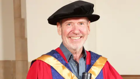 Steve Lillywhite, wearing a blue shirt underneath PhD robes and a hat, holds a degree scroll and smiles into the camera.