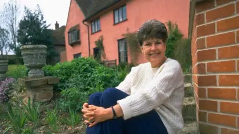 Getty Images A woman with short grey in front of a red coloured home. She sits on a step outside the home, resting her hands around her legs. She wears a white knitted jumper and blue jeans.