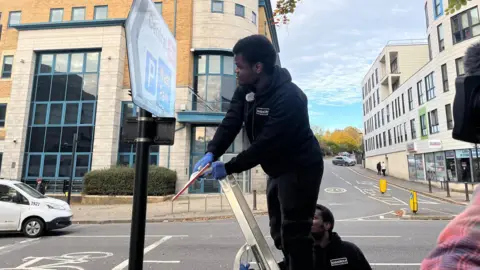 John Makanjuola stands on a ladder in a street in Southampton, holding a window wiper and cleaning a sign pointing towards the train station. He wears a black hoodie and black tracksuit while his twin brother can be seen holding the ladder wearing the same outfit.