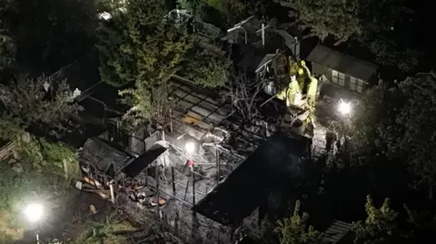 Charred remains for gardens seen at night with several firefighters among burnt out outbuildings.