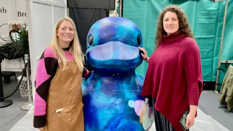Two women stand either side of a 1.5m high duck statue that has been decoratively painted in a marketplace entrance. The woman on the left has long blonde hair, a pink and black jumper, black leggings and boots. The duck is painted in blue and green swirls with white constellations and stars also painted on. The woman on the right has long dark curly hair, a long maroon jumper and black striped trousers. The duck is stood on a pallet and white ground sheets.