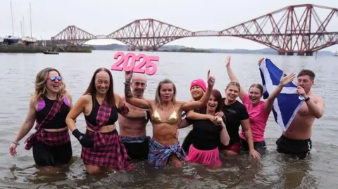 Nine people dressed in swimming costumes and tartan stand hip-deep in the Firth of Forth on a grey day. The Forth Rail Bridge can be seen in the background. 