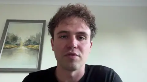 Matt Staniek during an online interview. He is looking at the camera. He is wearing a black T-shirt and has short messily styled hair. A painting of a lake hangs on the wall behind him.