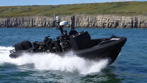 A black boat with two police officers travels quickly through the water near some cliff faces.