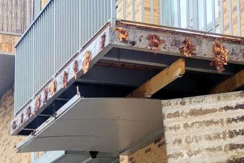 Close-up of a rusted steel balcony frame with exposed timber supports beneath a block of flats in daylight.