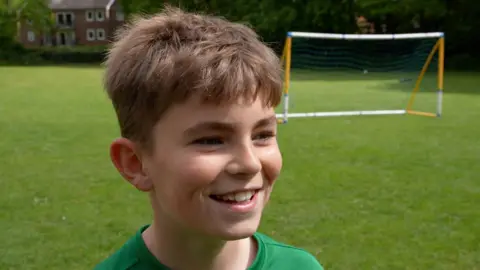 Ollie has short brown hair and stands on a grass pitch in front of a goal, smiling, in a green football top. 