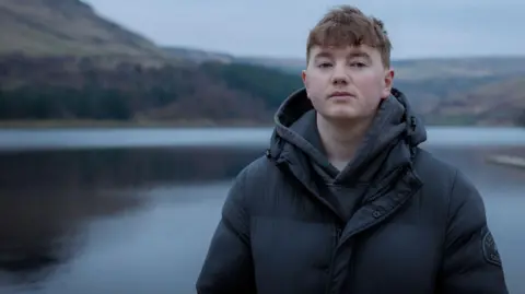 Alex Batty, who is wearing a black coat and hooded top, is standing in front of a large lake. 