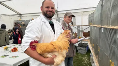The image shows a person in a white coat holding a large golden chicken inside a tent lined with cages, likely at a poultry show or judging event. Another person stands nearby with a clipboard, while additional chickens, cages filled with wood shavings, and a table with papers and flowers are visible in the background.