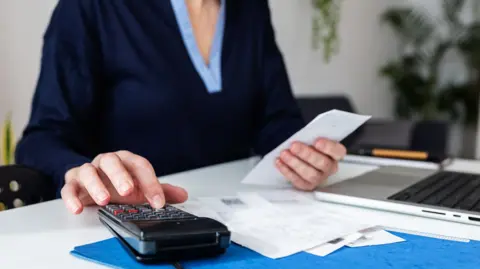 Woman wearing navy top with lighter blue trim. She is sitting at a desk with a laptop, some bits of paper and is using a calculator. There are some plants in the background