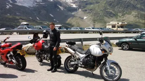 Gordon Castle Gordon Castle is wearing black leathers and is standing with three bikes around him and a snow-covered mountain side behind him. There is a road there too and several cars and a motorhome parked on the other side.