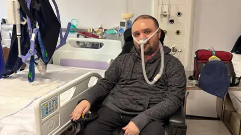 Erica Witherington / BBC Ravi Mehta is seated in a hospital chair beside a bed, wearing a nasal breathing tube connected to medical equipment. Surrounding items include a hoist sling, monitors, tubing and supplies, indicating a clinical setting with ongoing respiratory support.