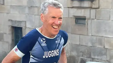Karen Juggins David Juggins, a man with short grey hair and wearing a blue Great Britain athletics outfit, smiles as he runs past during an event.