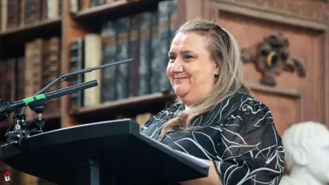 Jenny Magee Photographer Noeleen Timbery standing at a lectern in front of a microphone in the Wren Library, Trinity College, Cambridge