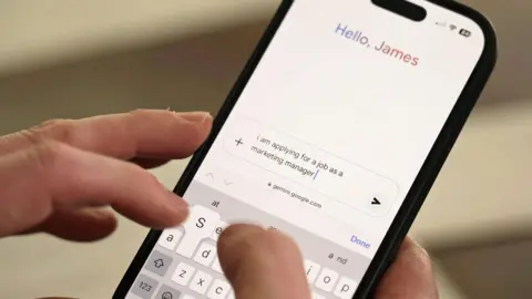 A close-up of a smartphone showing the Gemini chatbot interface. At the top of the phone a greeting reads "Hello, James" and a user is typing a prompt which begins "I am applying for a job as a marketing manager"