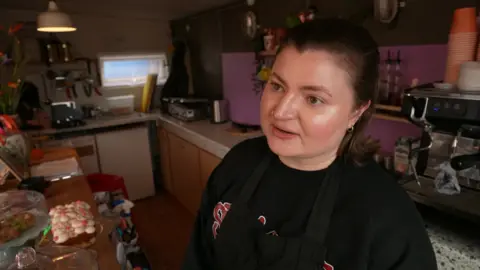 Alex Nixson-Evers is standing in the cafe kitchen in front of a counter filled with cakes.